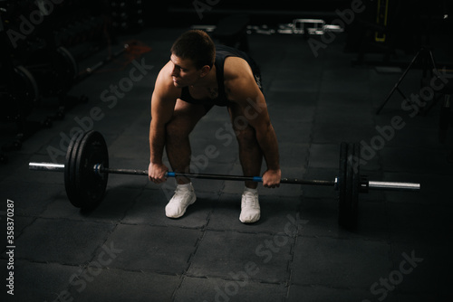 Muscular young man with perfect beautiful body wearing sportswear lifting heavy barbell from floor during sport workout training in modern dark gym. Concept of healthy lifestyle.