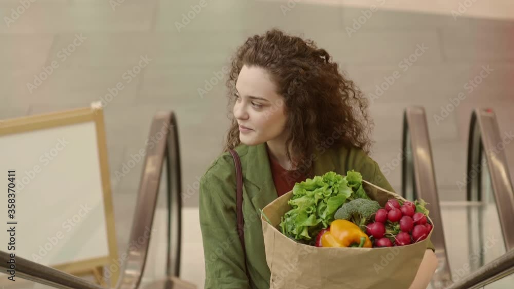 Medium shot of smiling curly-haired young woman wearing casual stylish clothes standing on escalator with food package in her hands Medium shot of smiling curly-haired young woman wearing casual stylish clothes standing on escalator with food package in her hands