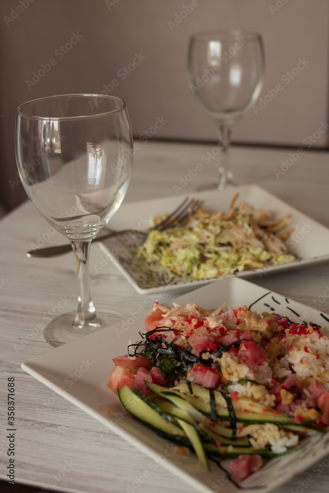 Beautiful glass transparent glasses and white square plates with food stand on a light wooden table
Beautiful glass transparent glasses and white square plates with food stand on a light wooden table