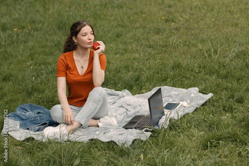Full lenght portrait of young dreamy girl with gadgets. Woman in orange t-shirt sitting on the green grass in park and eating red apple.