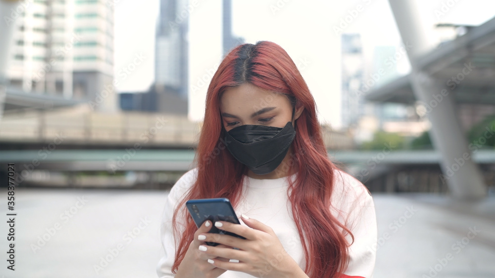 Obraz premium A woman dyed in red hair wearing a mask and standing playing with a mobile phone in the city. 4k Resolution.
