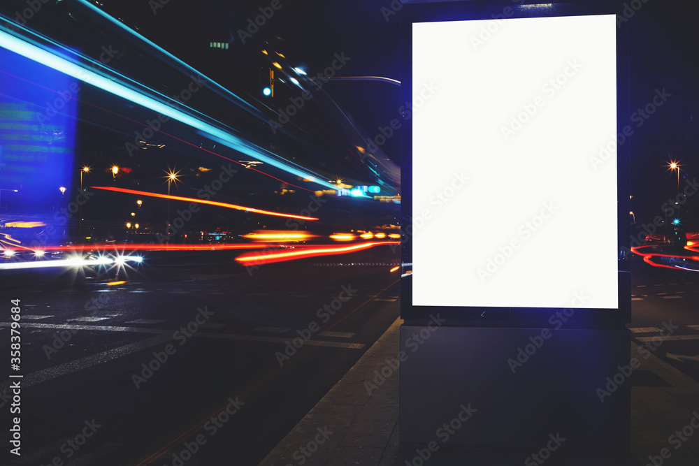 Illuminated blank billboard with copy space for your text message or content, public information board with shutter speed on background, advertising mock up in urban setting, empty poster on roadside