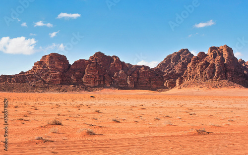 Red dusty desert with large rock massif and blue sky in background, small off road vehicle in foreground for scale. Typical landscape of Wadi Rum, Jordan