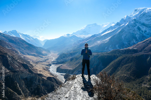 A man wearing a beanie and blue jumper, placing his hands together, breathing deeply the fresh mountain air. Freedom and happiness. Below Manang valley stretches in Himalayas, along Annapurna Circuit.