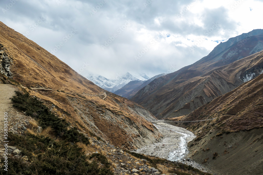 A view on rushing brook in Manang Valley, Annapurna Circus Trek, Himalayas, Nepal, with  Annapurna Chain and Gangapurna in the back. Dry and desolated landscape. High, snow capped mountain peaks.