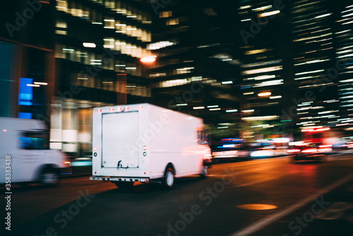 White truck of logistic company driving on city road delivering goods for order quickly in time, truck moving speedly on avenue transporting and distributing stuff for postal service in megalopolis photo