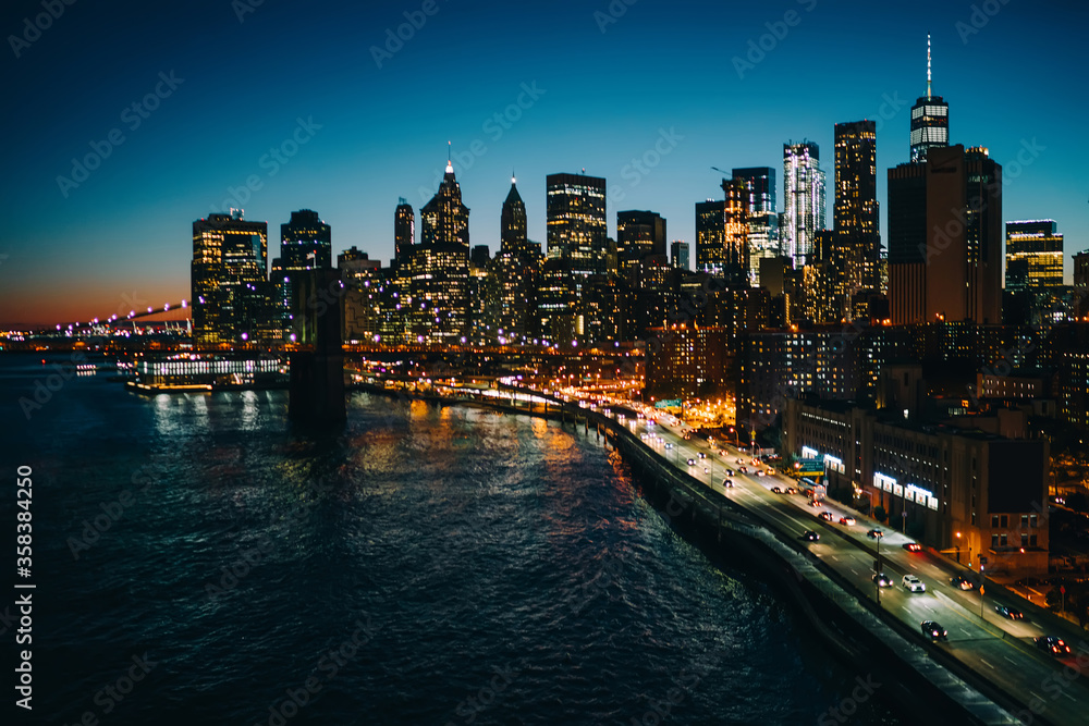 Panoramic view tall skyscrapers buildings near river with real estate luxury apartments illuminating at night, developed infrastructure of New York megalopolis with traffic and modern architecture