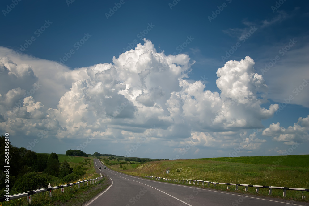 Long road. Highway among white clouds in a blue sky. summer landscape green trees and grass