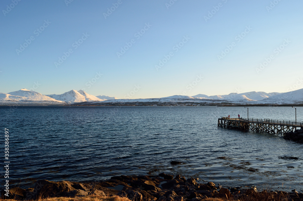 sunshine over blue fjord and pier with snowy mountain background