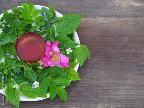 Herbal tea in a glass cup and medicinal aromatic plants - raspberry leaves, black currant, mint, rose hips, strawberry flowers, glechoma hederacea in a white plate on a wooden stand, top view,