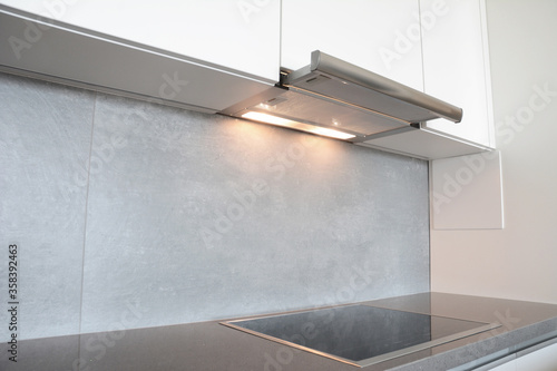 A close-up on under cabinet range hood, exhaust vent hood with lights working and modern electric stove, cooktop in gray and white kitchen design.