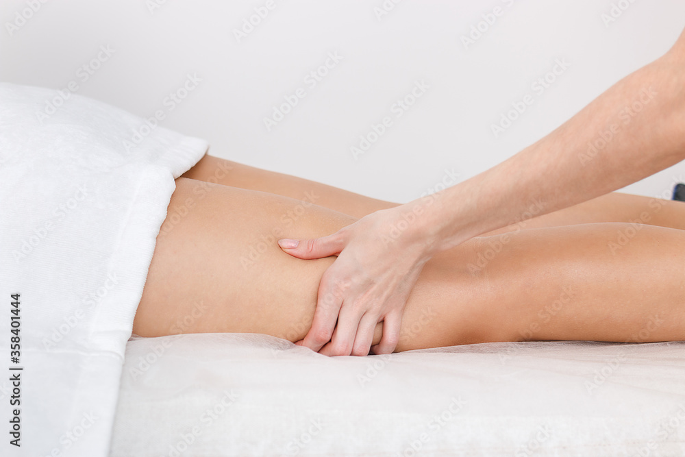 Kneading with a massage of the muscles of the legs on the hips of a model lying on a couch against a white wall.