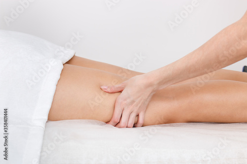 Kneading with a massage of the muscles of the legs on the hips of a model lying on a couch against a white wall.
