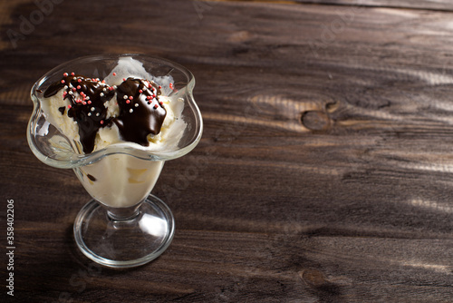 White balls of vanilla ice cream poured with chocolate topping in a dessert glass. Decorated ice cream on an old table made of rough wood. Copyspace.