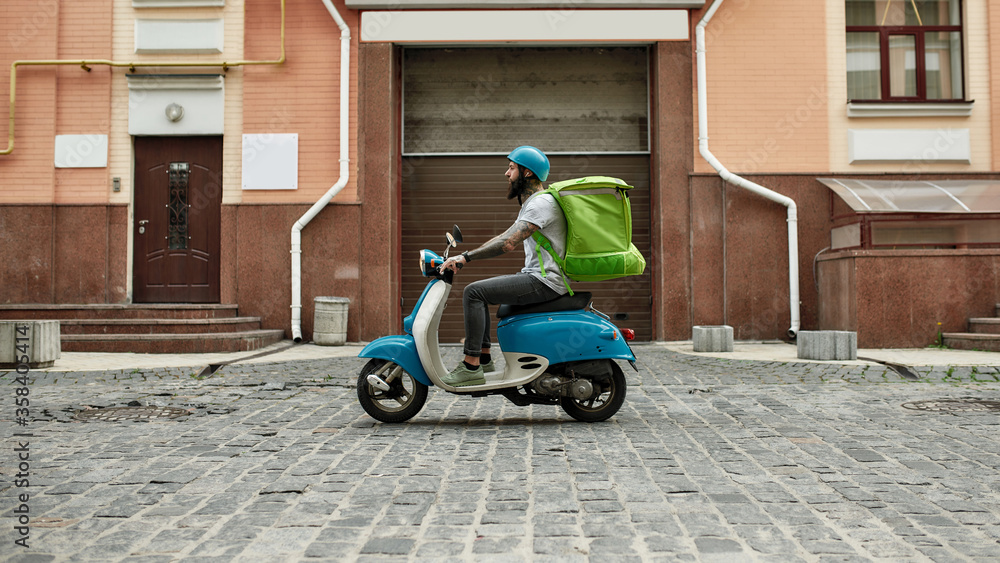Trendy Food Delivery. Bearded delivery man in helmet with thermo bag or backpack riding a motor scooter along the city, delivering food. Courier, delivery service concept