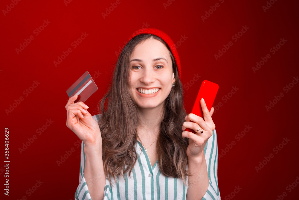 Excited young woman is holding a phone and credit or debit card over red background Excited young woman is holding a phone and credit or debit card over red background