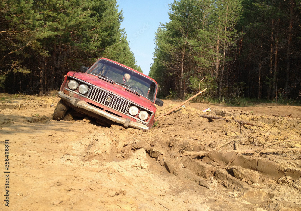 A red passenger car stuck deep in the mud on a forest road.