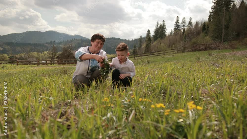Grandmother makes wreath of wild flowers with her young grandson. Who wears traditional costume embroidery shirt, family making homemade, I love my gran. Relax outside on green grass, behind mountain Grandmother makes wreath of wild flowers with her young grandson. Who wears traditional costume embroidery shirt, family making homemade, I love my gran. Relax outside on green grass, behind mountain