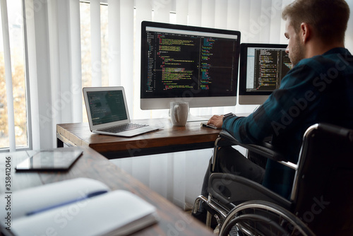 I can work with any code. Concentrated male web developer in a wheelchair writing program code on multiple computer screens while sitting at his workplace in the modern office