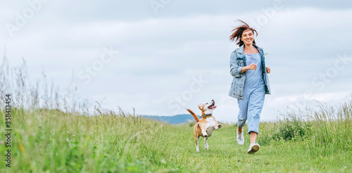 Happy smiling jogging female with fluttering hairs and her beagle dog running and looking at eyes. Walking by meadow grass path in nature with pets, healthy active people lifestyle concept image.