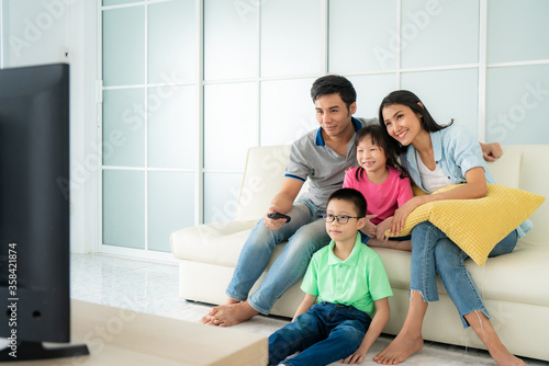 Asian happy family with father, mother, son and daughter sitting in sofa and watching television at home concept of family values, vacation day, holiday, happiness or lifestyle.