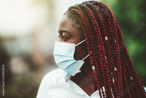 Close-up portrait of a young African woman with chestnut braids and in a virus protective mask over her face; masked black woman outdoors - protection against influenza and pandemic, selective focus photo