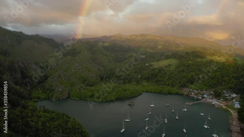 Aerial tilt up shot of harbor by village near rainbow against cloudy sky, drone flying over boats moored on sea near green mountains - Wakatobi Regency, Indonesia