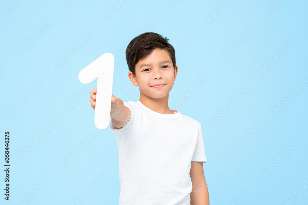 Portrait of smiling young Asian boy holding and showing number one cutout in isolated studio blue background for education concept Portrait of smiling young Asian boy holding and showing number one cutout in isolated studio blue background for education concept