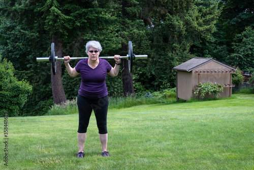 Middle aged caucasian woman with gray hair lifting a barbell with black plates, fitness outside on the lawn  © knelson20