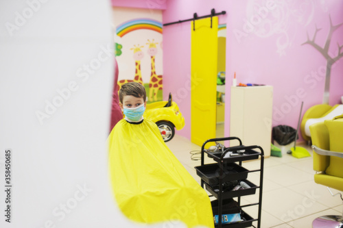 preschool boy in protective mask with yellow Cape on body is sitting in Barbers chair. work of beauty salons compliance with safety rules, children haircuts in compliance with protective measures photo