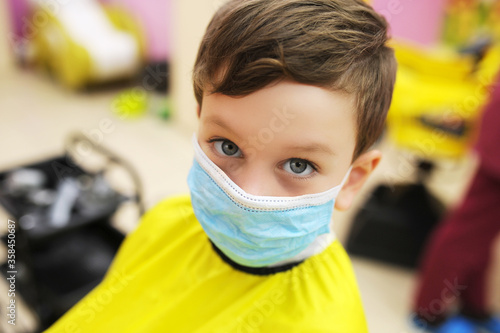 preschool boy in protective mask with yellow Cape on body is sitting in Barbers chair. work of beauty salons compliance with safety rules, children haircuts in compliance with protective measures photo