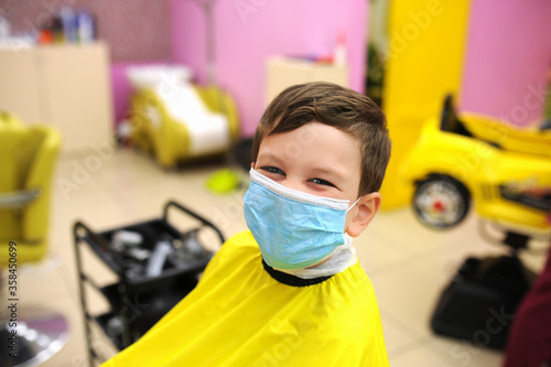 preschool boy in protective mask with yellow Cape on body is sitting in Barbers chair. work of beauty salons compliance with safety rules, children haircuts in compliance with protective measures photo