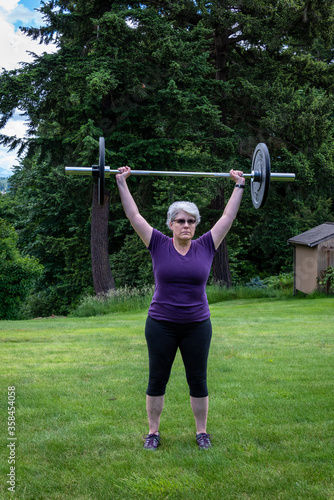 Middle aged caucasian woman with gray hair lifting a barbell with black plates, fitness outside on the lawn 