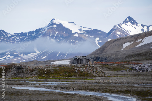 Abandoned wooden cabin and red bricks on the ground. Snow covered mountains in the background and melting water streams in the foreground. © Sandra