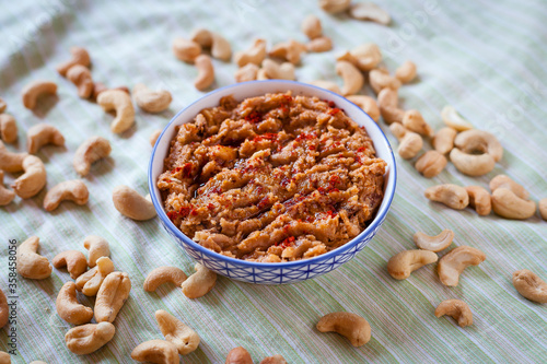 Food plated shot of a small blue and white bowl with vegan sobrasada or cashew dip on a green striped tablecloth with dry tomato, paprika, olive oil, garlic and pepper and surrounded by  dry cashews.