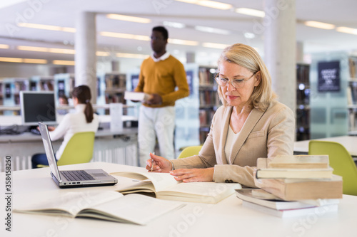 Mature confident woman with laptop and book in public library