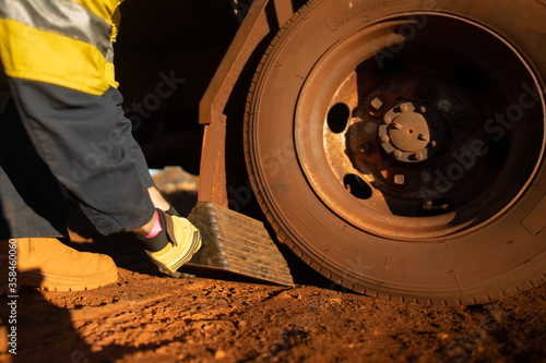 Safety workplaces close up picture of miner track driver wearing safety hand glove placing wheel chock under back wheel to stable preventing its moving, rolling forward or backward construction site 