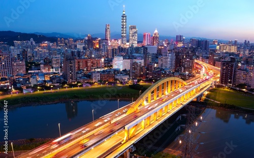 Night skyline of Taipei City viewed from above a riverside park with a highway bridge spanning Keelung River & towers standing out amid the skyscrapers in Xinyi Commercial District under sunset sky