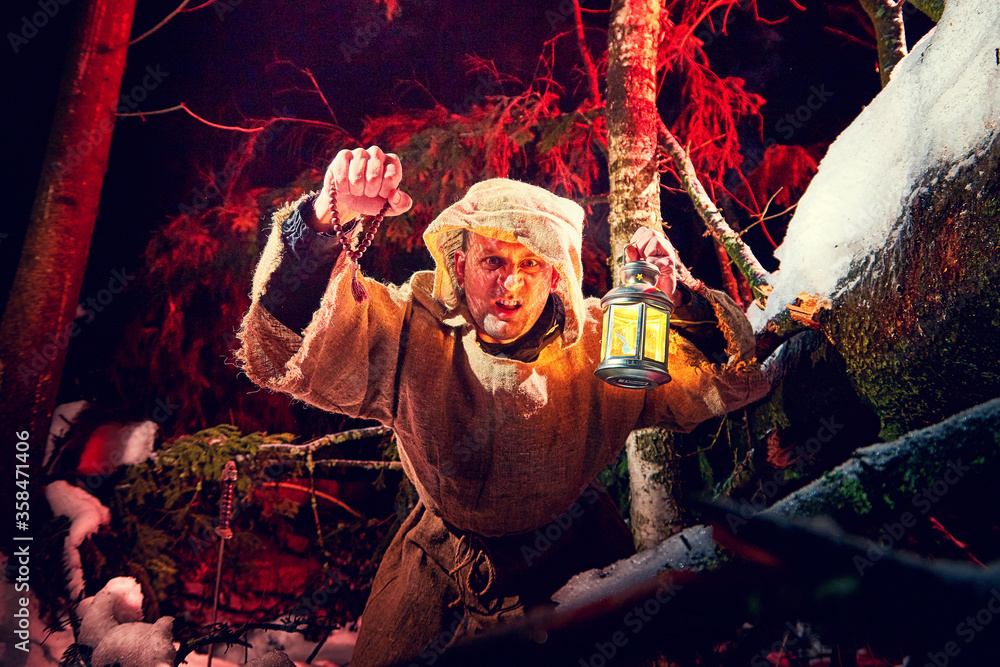 Obraz premium Evil medieval monk in canvas sackcloth robe with lattern in dark forest with snow and red light on winter night. Fantasy or fairy tale about wandering monk. Forces of good and evil in world