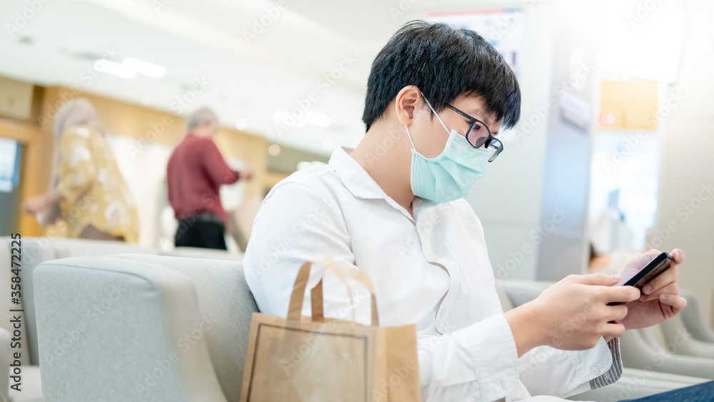 Fototapeta premium Male Asian patient wearing surgical face mask using smartphone at waiting area in hospital or medical centre. Medical exam or body check up. Coronavirus pandemic prevention. Health care concept