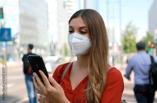 Business woman wearing KN95 FFP2 face mask walking in modern city street holding a smartphone with people on the background