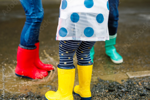 Close-up of three children, toddler girl and two kids boys wearing red, yellow and green rain boots and walking during sleet. Happy siblings jumping into puddle. Having fun outdoors, active family