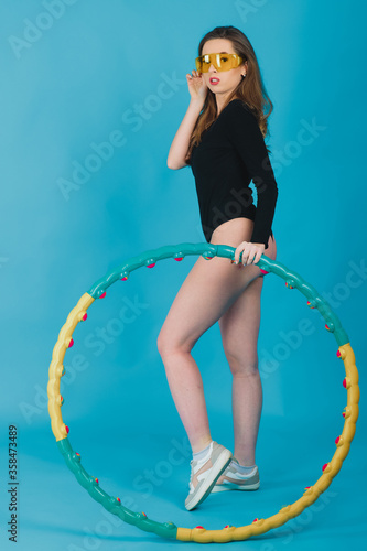 beautiful sports girl dressed in black bodysuit in yellow glasses with hula hoop on blue background