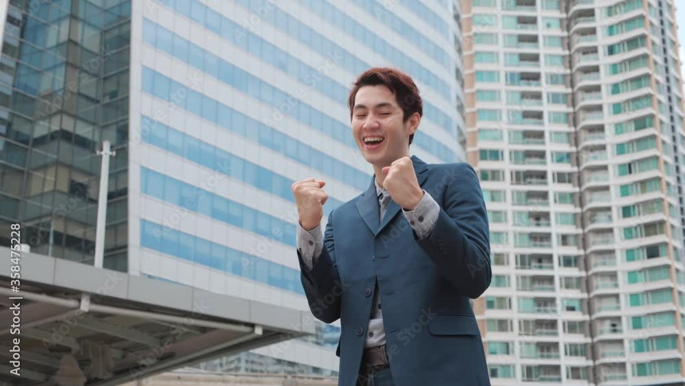 Asian Business win. Asian Businessman look at the camera shaking fists celebrating success at work with modern office building background. business success, achievement, and win concepts Asian Business win. Asian Businessman look at the camera shaking fists celebrating success at work with modern office building background. business success, achievement, and win concepts