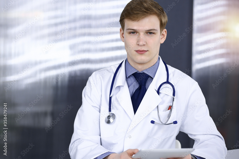 Blond male doctor sitting and working with tablet computer in sunny clinic at his working place, close-up. Medicine concept Blond male doctor sitting and working with tablet computer in sunny clinic at his working place, close-up. Medicine concept