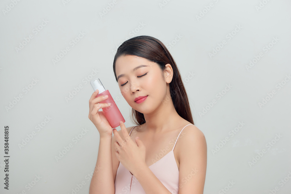 Young pretty stylish woman over white wall background holding bottle with cosmetics water. Young pretty stylish woman over white wall background holding bottle with cosmetics water.