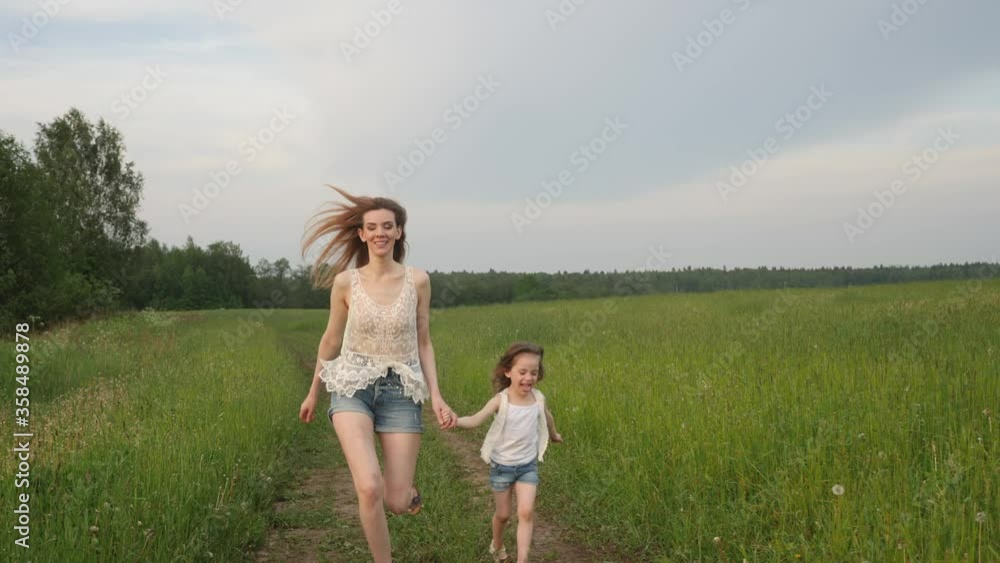 young mother in short shorts run with her daughter a child at sunset on a green field along the road young mother in short shorts run with her daughter a child at sunset on a green field along the road