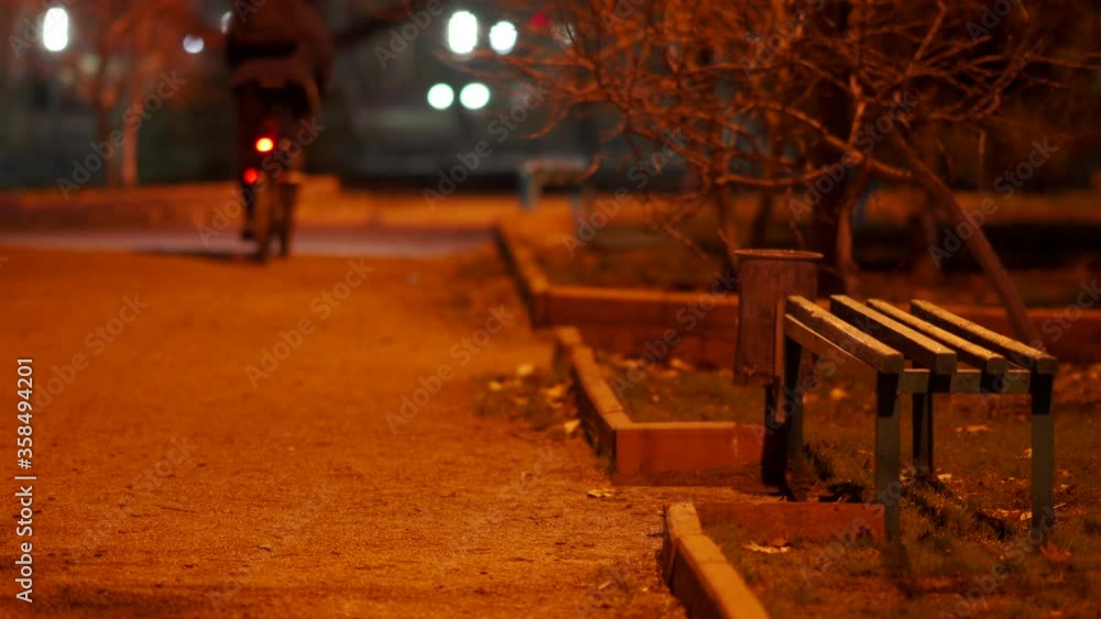 Cyclist riding a bike along empty dark footpath at night, street lights illuminate a lonely alley in the fall.