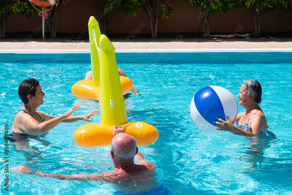 Happy retired couples having fun in swimming pool with volleyball in the water - four senior friends or family in active elderly vacation