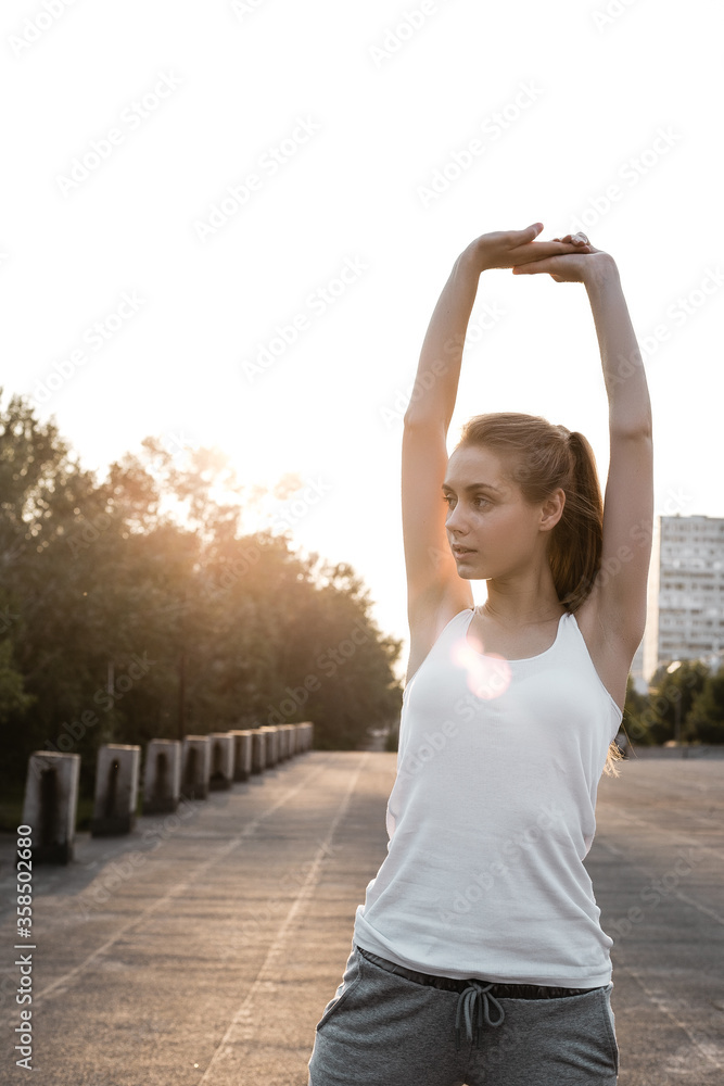 Girl stretching her hands outdoor and preparing to sport trainings. Female model posing outside on air in sporty clothes and doing exercises
